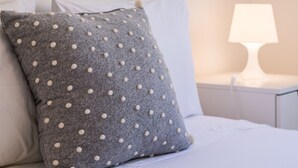 A neatly made bed with a grey polka-dotted pillow and white pillows, a white bedside table with a lamp, and a white wall. - LAGOS CLASSIC by HOMING (Lagos)