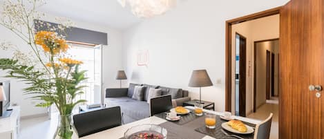 A modern hotel room with a dining area, a sofa, a laptop, and a breakfast setup.
