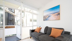 Een moderne woonkamer met een grijze bank, twee oranje kussens, een groot raam en een schilderij van een zandstrand aan de muur.