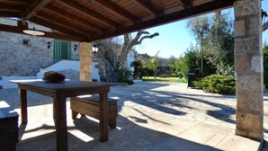 Un espace extérieur aménagé en terrasse, avec une table et des bancs en bois, un sol en pierre et un mur en pierre agrémenté de volets verts.