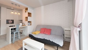 Un espace de vie compact comprenant un coin cuisine, un canapé gris agrémenté de coussins rouges, une table à manger blanche et un petit espace cuisine avec des luminaires suspendus.
