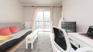 Une pièce moderne et bien éclairée, avec un canapé gris, un bureau blanc, une télévision et une grande fenêtre avec des rideaux.