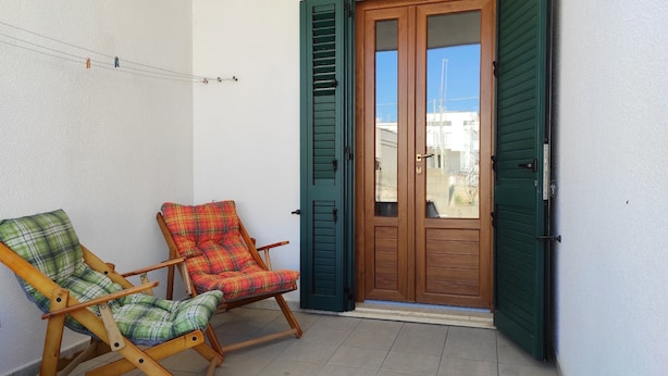 Un piccolo balcone con due sedie di legno, una porta in legno con persiane verdi e vista sugli edifici.