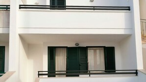 A modern two-story building with a flat roof, a balcony with green shutters, and a staircase leading to the upper floor.