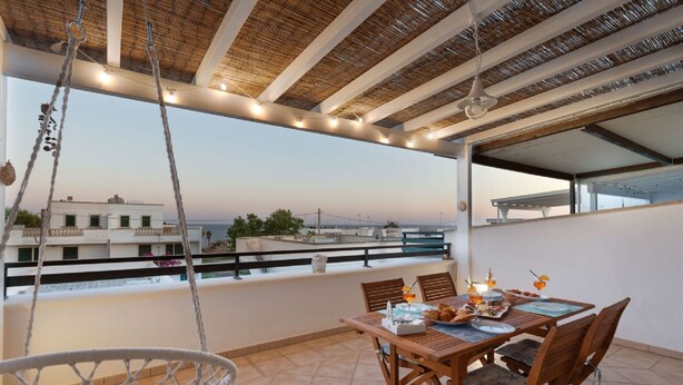 A balcony with a wooden table set for dining, a hanging chair, and a view of the cityscape.