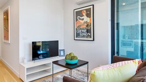 Una sala moderna con un televisor de pantalla plana sobre un soporte blanco, una mesita con un cuenco azul y un cartel en la pared.