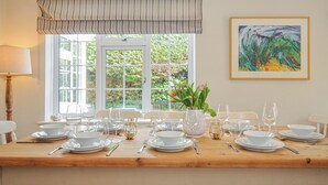 Una mesa de comedor preparada para seis personas, con platos, tazones y vasos blancos, un jarrón con flores y un cuadro en la pared.