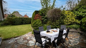 Un patio con una mesa y sillas de piedra, rodeado de abundante vegetación y un cielo azul despejado.