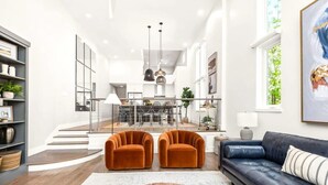 Un salon moderne avec un canapé bleu foncé, deux fauteuils orange et une table basse en bois. Un escalier mène à une salle à manger équipée d’une table et de chaises en verre.