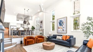 Un vaste espace de vie agrémenté d’un canapé bleu foncé, de fauteuils orange et d’une table basse en bois. Un grand tableau abstrait orne le mur, et la pièce est dotée d’un plafond haut avec un ventilateur de plafond.