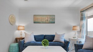 Una sala de estar con un sofá azul, una mesa de centro blanca con una planta verde, y un cuadro de veleros en la pared.