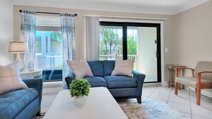 Un salón con un sofá azul, una mesa de centro blanca y una planta en maceta. Hay un ventilador de techo, una lámpara y vistas al exterior a través de una puerta corrediza de vidrio.