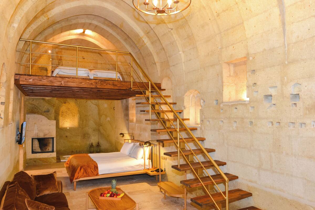 A room with a bed, a wooden bench, a small table with fruit, a sofa, and a staircase.