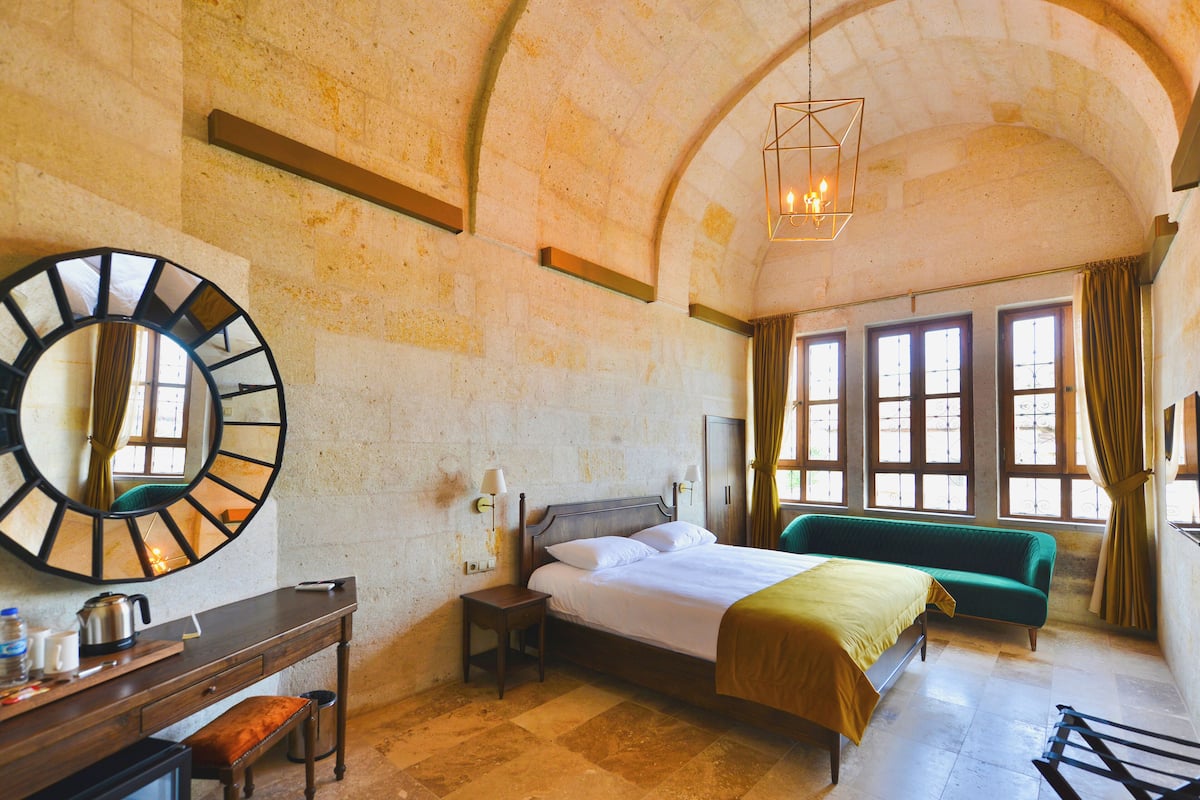 A hotel room with a large mirror, a bed with a green bedspread, a desk with a chair, and a hanging light fixture.