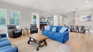 Amplia sala de estar con un sofá azul, una mesa de centro de madera y un sillón reclinable de cuero negro. Se ve una cocina al fondo, con electrodomésticos modernos y una zona de comedor con mesa y sillas de madera.