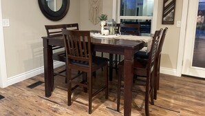 A dining area with a wooden table and chairs, a round mirror, a macrame wall hanging, and a window with a view of the outside.