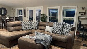 A brown leather sectional sofa with patterned pillows and a throw blanket, a dining table with chairs, and a potted plant.