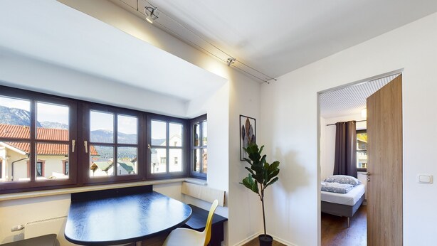 Una habitación moderna con una mesa de comedor, dos sillas, una planta en maceta y una cama en la habitación contigua.