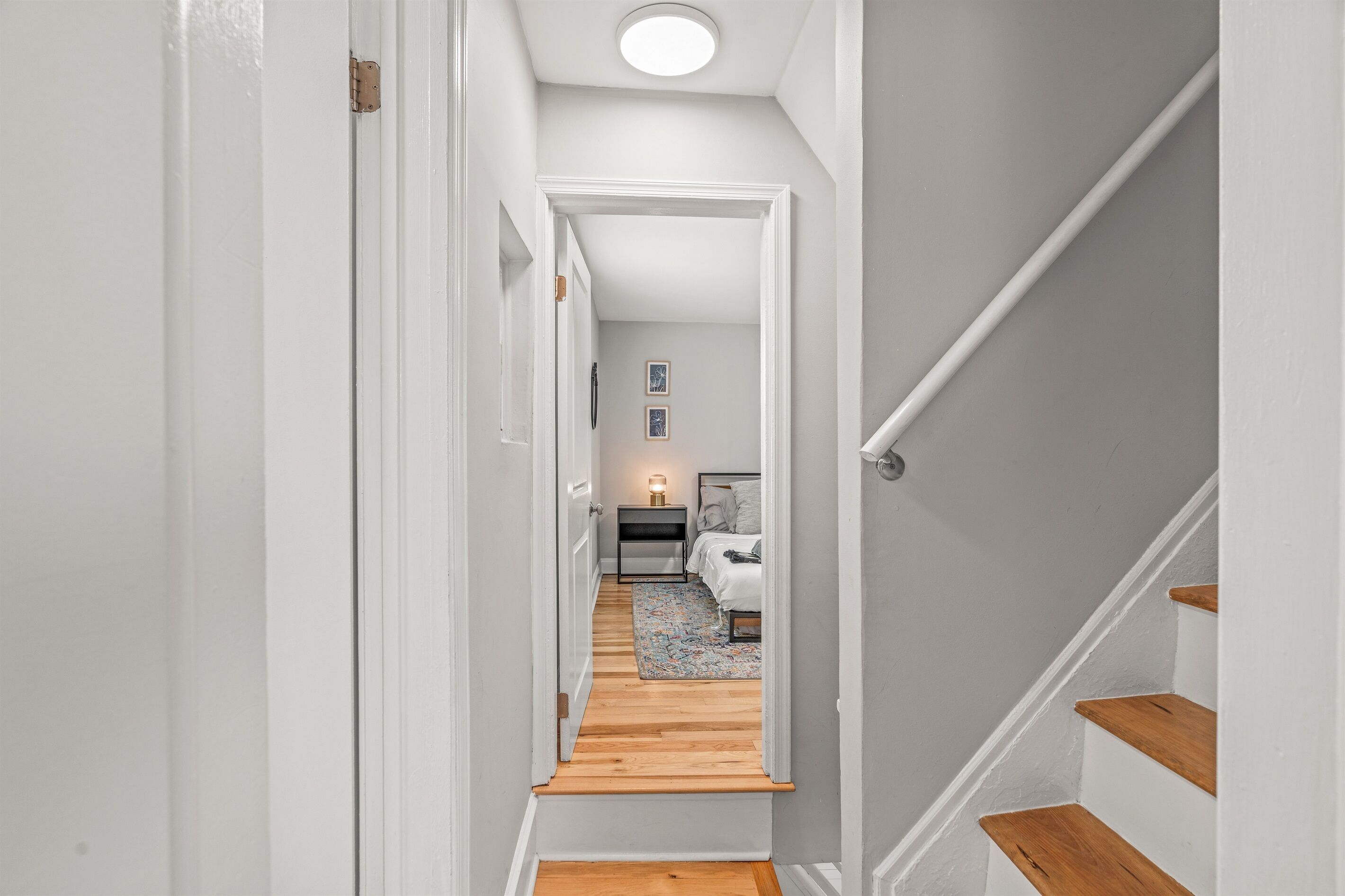 Un couloir étroit, doté d’un plancher en bois, mène à une chambre à coucher qui contient un lit, une table de chevet et une petite table de nuit avec une lampe.