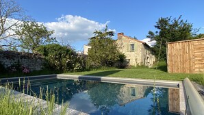Una piscina rectangular con borde de madera, rodeada de abundante vegetación y un muro de piedra.