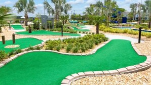 Ein Minigolfplatz mit grünem Kunstrasen, umgeben von Palmen und einem Poolbereich.