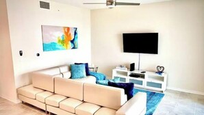 Ein Wohnzimmer mit einem beigefarbenen Sofa, einem Flachbildfernseher, einem Deckenventilator und einem farbenfrohen abstrakten Gemälde an der Wand.
