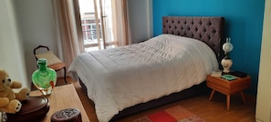 Une chambre à coucher avec un lit, une table de chevet, une lampe, un ours en peluche, une bouteille et une fenêtre avec des rideaux.