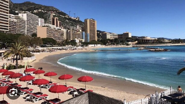 Un hotel frente a la playa con sombrillas rojas, arena y vistas al mar y a las montañas.