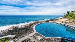 A pool with a curved edge, surrounded by a stone deck and a railing, overlooking a rocky shoreline and the ocean. - Casa De Emdeko 210 (Kailua-Kona)