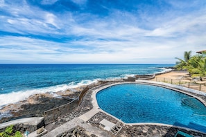 Ein Pool mit gewölbter Kante, umgeben von einer Steindeck und einer Geländer, mit Blick auf eine felsige Küstenlinie und den Ozean.