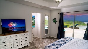 Un dormitorio con una cama grande, un aparador, un televisor mostrando fuegos artificiales, un ventanal corredero a un balcón y vistas a la playa.