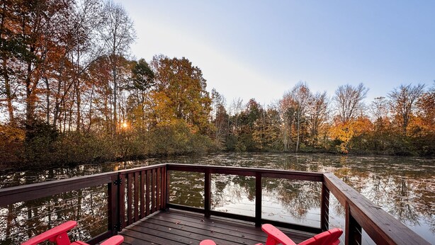 A wooden deck with two red Adirondack chairs overlooking a calm body of water surrounded by trees with autumn foliage. - The 'Happy Go Fishing Cabin' at the Park Place (on Parks Rd)  (Woodleaf)