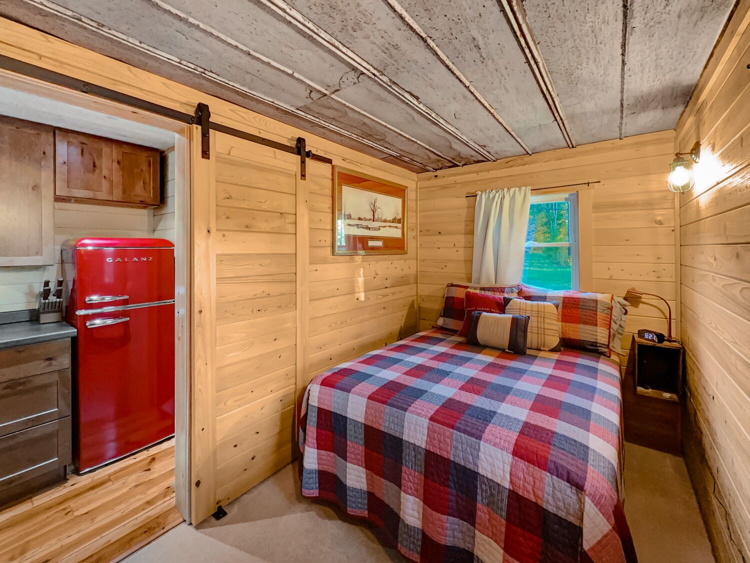 Une chambre confortable, aménagée dans le style d’un chalet, avec un lit, un réfrigérateur Galanz rouge, des murs en bois et une fenêtre avec des rideaux.