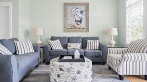 A living room with a grey sofa set, striped pillows, a round ottoman with nautical prints, and a framed seagull painting on the wall.