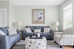 Un salon avec un canapé gris, des coussins à rayures, un pouf rond orné de motifs marins et, au mur, un tableau représentant une mouette.