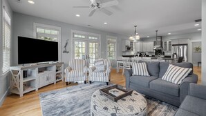 A spacious living room with a large flat-screen TV, striped armchairs, a grey sofa, and a round ottoman with a book.