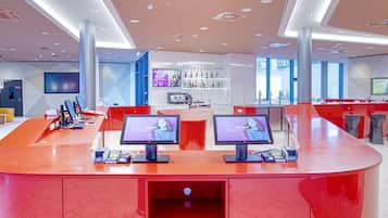 A modern hotel reception area with a curved red counter, two monitors displaying welcome messages, a bar area with bottles, and a large TV screen.