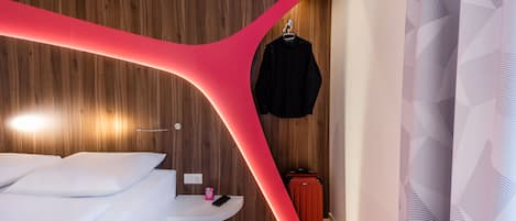 A modern hotel room with a wooden wall, a white bed, a red accent wall, a hanging black jacket, and a suitcase.