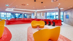 A modern lobby with a red reception desk, yellow and orange seating, and large windows offering a view of the city. - Prize by Radisson, Vienna City (Vienna)