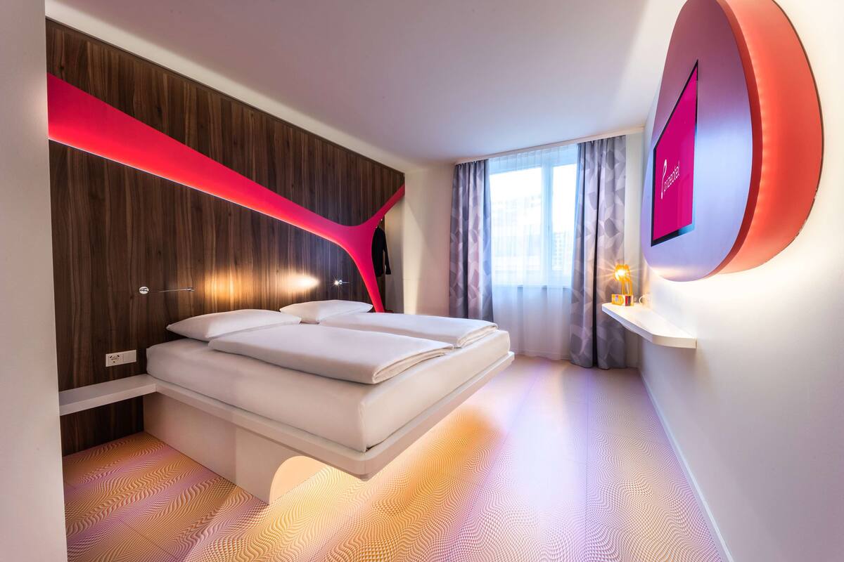 A modern hotel room with a bed, a wall-mounted TV, and a window with curtains.