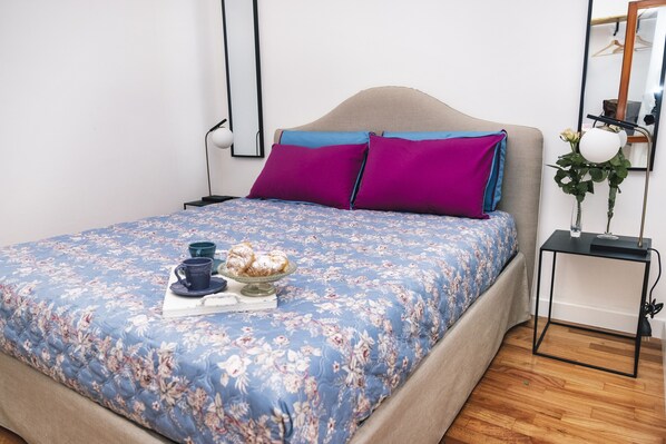 En pænt redt seng med blomstret sengetæppe, to puder, et sengebord med lampe og en lille vase med blomster.