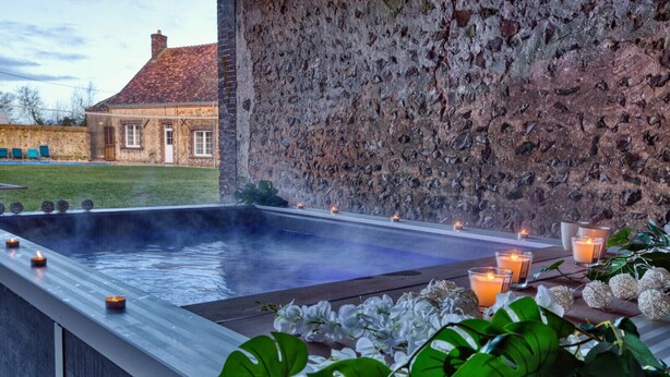 Een bubbelbad met kaarsen en planten, voor een stenen muur en een traditioneel gebouw op de achtergrond.