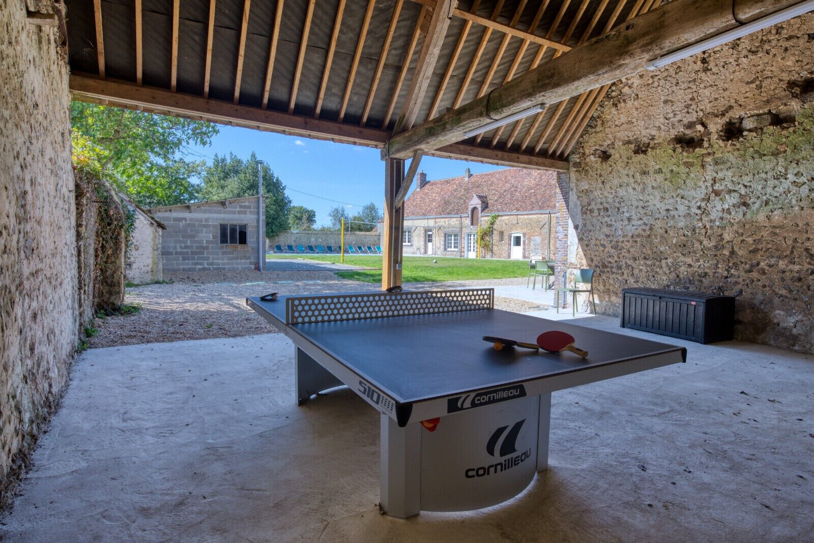 A ping pong table with a ball and paddle in a covered outdoor area with stone walls and a building in the background.
