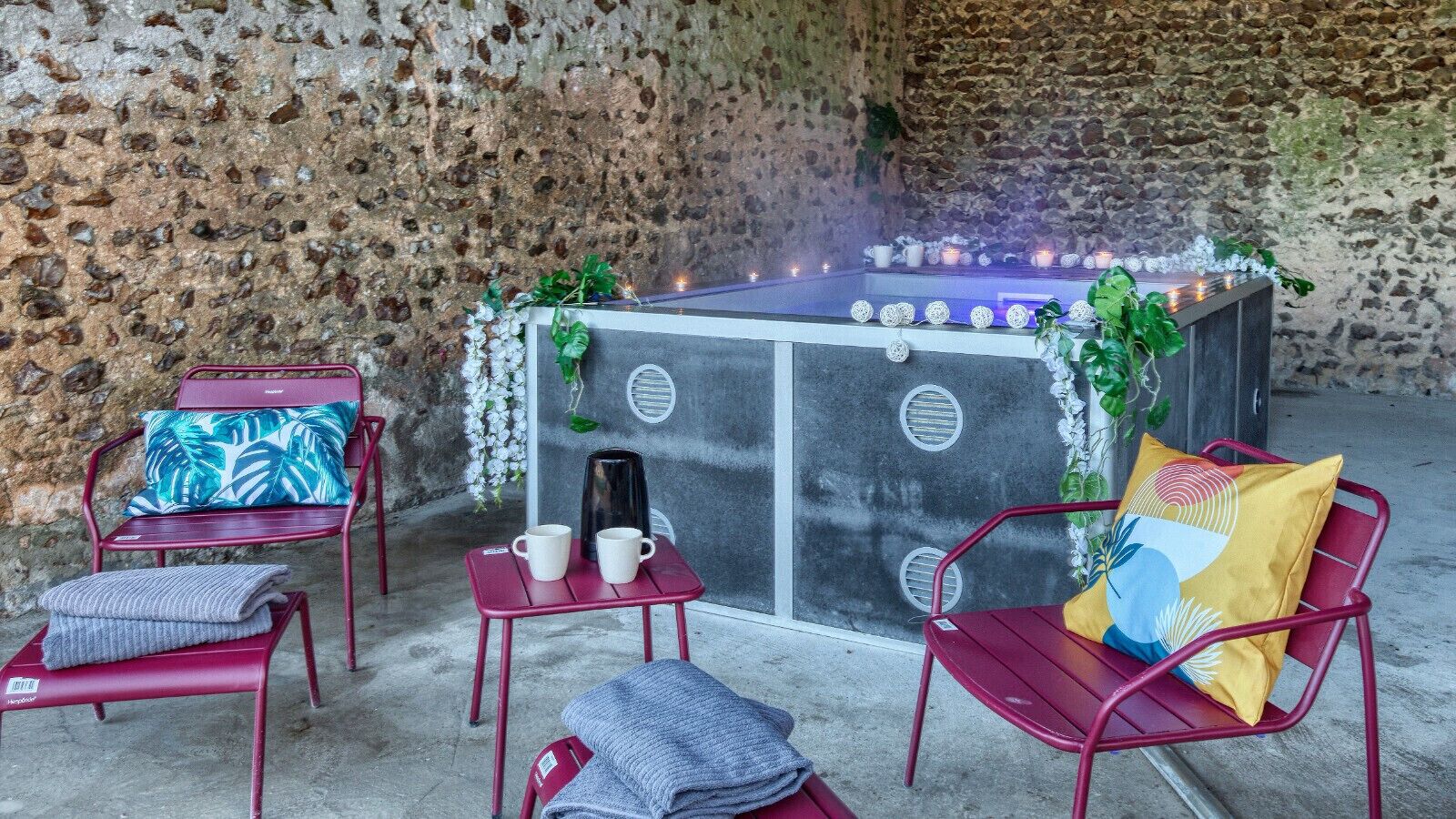 A cozy outdoor seating area with a stone wall backdrop, featuring a central table with candles, surrounded by red metal chairs and a small table with two cups.