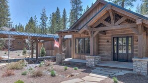A log cabin with a stone foundation, a covered porch, and a flag. - Guest Fav! Pool table, ping pong, foosball, paddle boards, AC, Hot tub, pet friendly, luxury home, family friendly, this home has it all! (Sunriver)