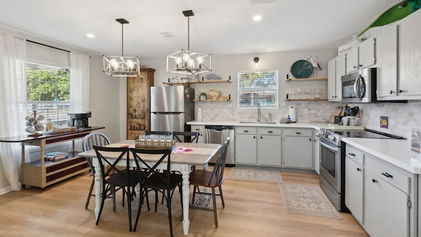Una cocina moderna con armarios blancos, una zona de comedor con mesa y sillas de madera, y un amplio ventanal con cortinas.