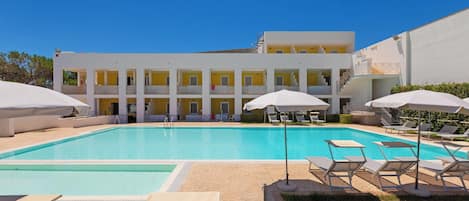 A swimming pool with lounge chairs and umbrellas, a building with multiple balconies, and a clear blue sky.