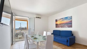 A modern living room with a glass dining table, white chairs, a blue sofa, and a sunset painting on the wall.