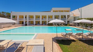 Una piscina con sillones y sombrillas, un edificio con varios balcones, y un cielo azul despejado.