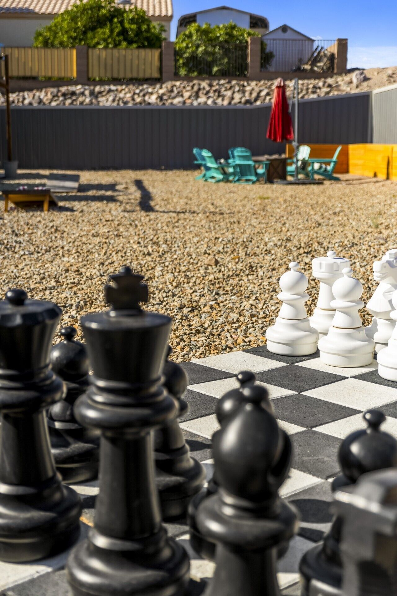 A chessboard with black and white pieces, a gravel surface, and a fence in the background.